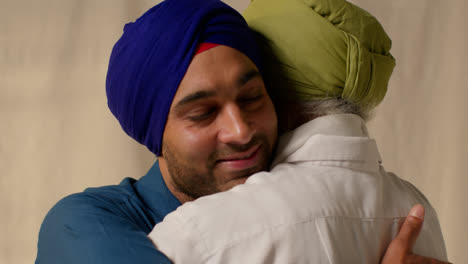 Close-Up-Studio-Shot-Of-Adult-Sikh-Son-Embracing-Senior-Father-Both-Wearing-Turbans-Against-Plain-Background-1